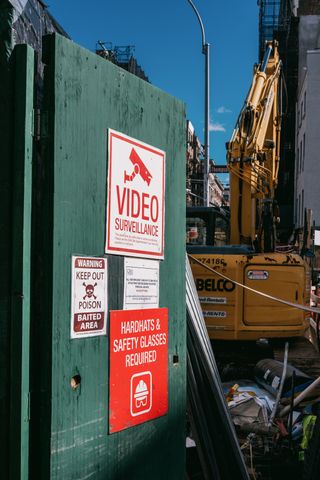 A partially open temporary green door covered with warning signs at a construction site, with an excavator visible in the background