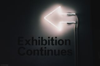 A neon sign on a wall in the shape of an arrow pointing left, above the words “Exhibition Continues”